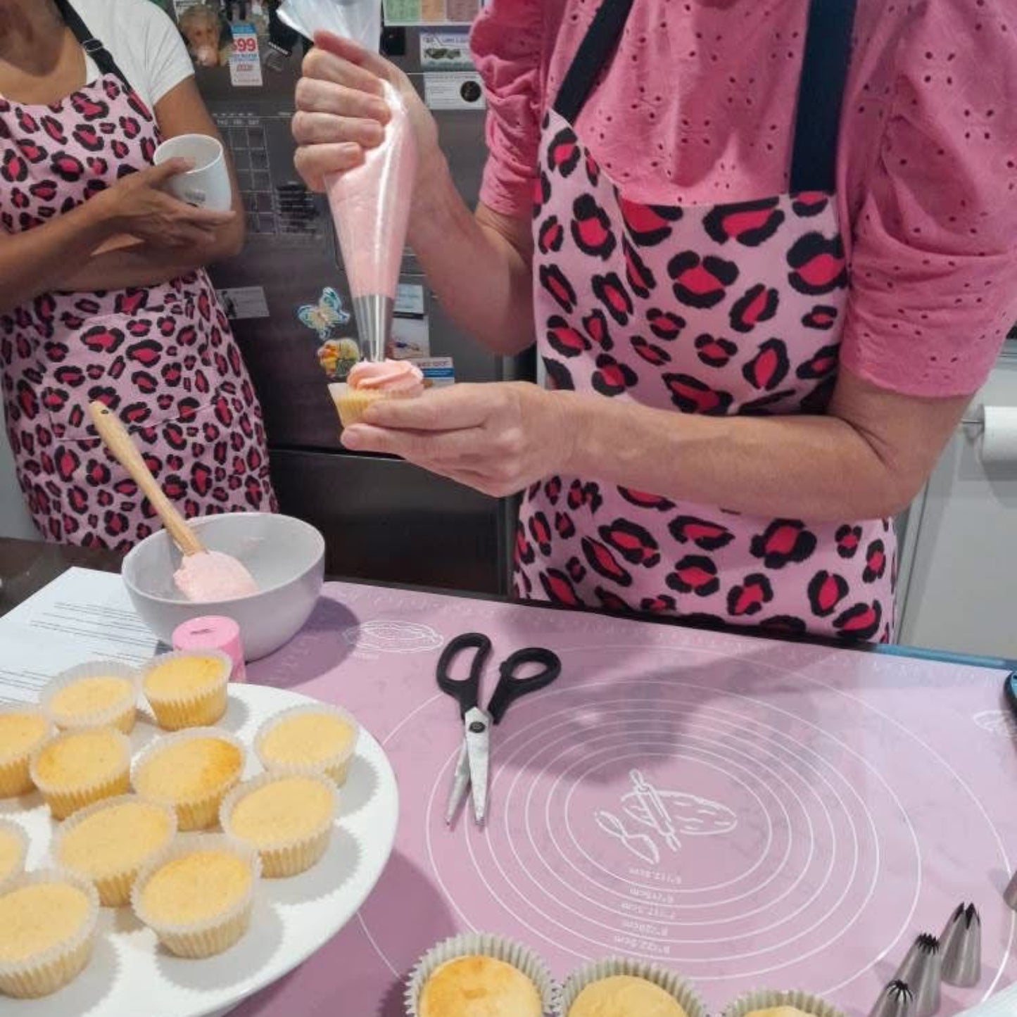 Leesa from Sweet As Co. in pink aprons with leopard print patterns decorating cupcakes in cupcake decorating class.