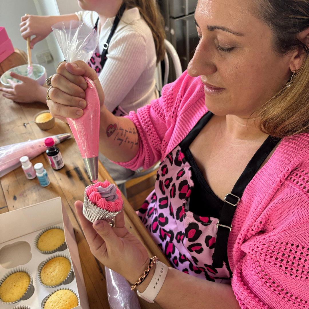 Woman in pink sweater decorating a cupcake with pink buttercream frosting in a kitchen in cupcake decorating class.