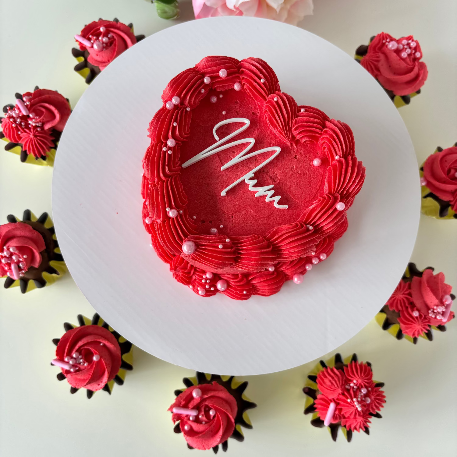 Red 4 inch heart-shaped cake with 'Mum' acrylic topper on a white plate surrounded by red cupcakes.